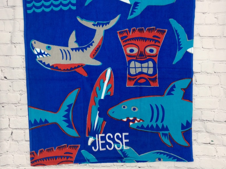 May include: Blue beach towel with shark, surfboard, and tiki designs. The towel features a shark with gray and red accents, a surfboard with red and blue accents, and a tiki with red and silver accents. The name "JESSE" is embroidered in white.