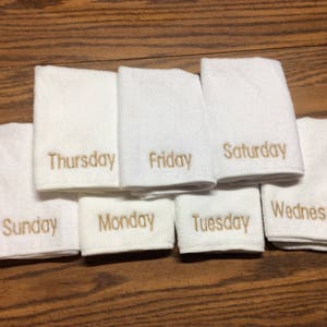 May include: White hand towels embroidered with the days of the week in gold thread. The towels are arranged on a wooden surface. Each towel is labeled with a day: Sunday, Monday, Tuesday, Wednesday, Thursday, Friday, and Saturday.