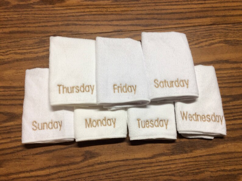 May include: White hand towels embroidered with the days of the week in gold thread. The towels are arranged on a wooden surface. Each towel is labeled with a day: Sunday, Monday, Tuesday, Wednesday, Thursday, Friday, and Saturday.