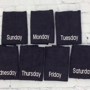 May include: Seven black washcloths, each embroidered with a different day of the week in white script: Sunday, Monday, Tuesday, Wednesday, Thursday, Friday, and Saturday. They are arranged against a white brick wall.
