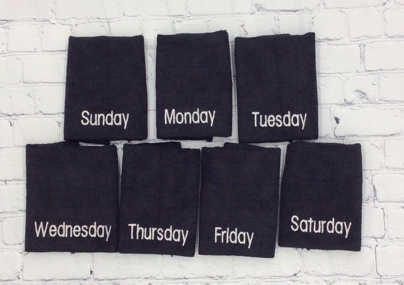 May include: Seven black washcloths, each embroidered with a different day of the week in white script: Sunday, Monday, Tuesday, Wednesday, Thursday, Friday, and Saturday. They are arranged against a white brick wall.