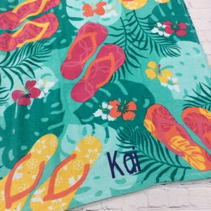 May include: A turquoise beach towel with a tropical design featuring colorful flip-flops, hibiscus flowers, and palm leaves. The name "Kai" is embroidered in blue. The towel is perfect for a day at the beach or pool.