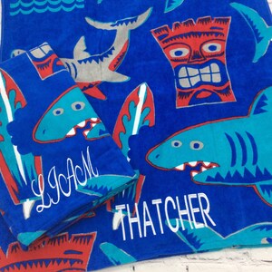 May include: Blue beach towels with shark, surfboard, and tiki designs. One towel is folded, displaying the name "LIAM" in white script. The other towel shows the name "THATCHER". The towels have a fun, summery theme.