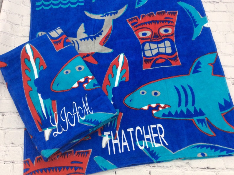 May include: Blue beach towels with shark, surfboard, and tiki designs. One towel is folded, displaying the name "LIAM" in white script. The other towel shows the name "THATCHER". The towels have a fun, summery theme.