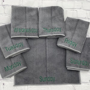 May include: A set of seven grey microfiber cleaning cloths, each embroidered with a day of the week in green lettering. The cloths are arranged on a white brick background.