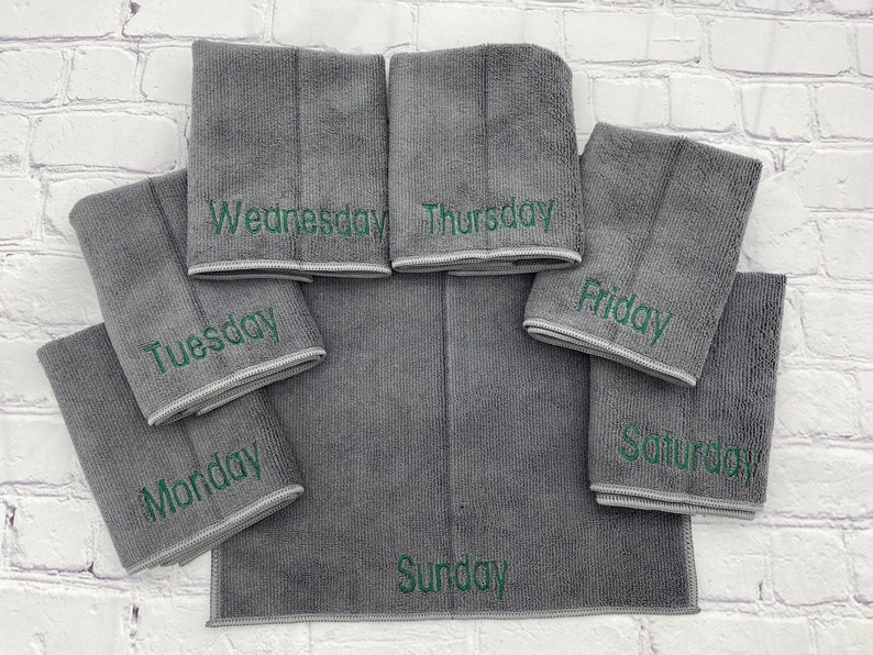 May include: A set of seven grey microfiber cleaning cloths, each embroidered with a day of the week in green lettering. The cloths are arranged on a white brick background.