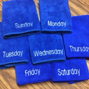 May include: Seven blue microfiber cloths, each embroidered with a day of the week in white. The cloths are arranged on a wooden surface. The days of the week are Sunday, Monday, Tuesday, Wednesday, Thursday, Friday, and Saturday.