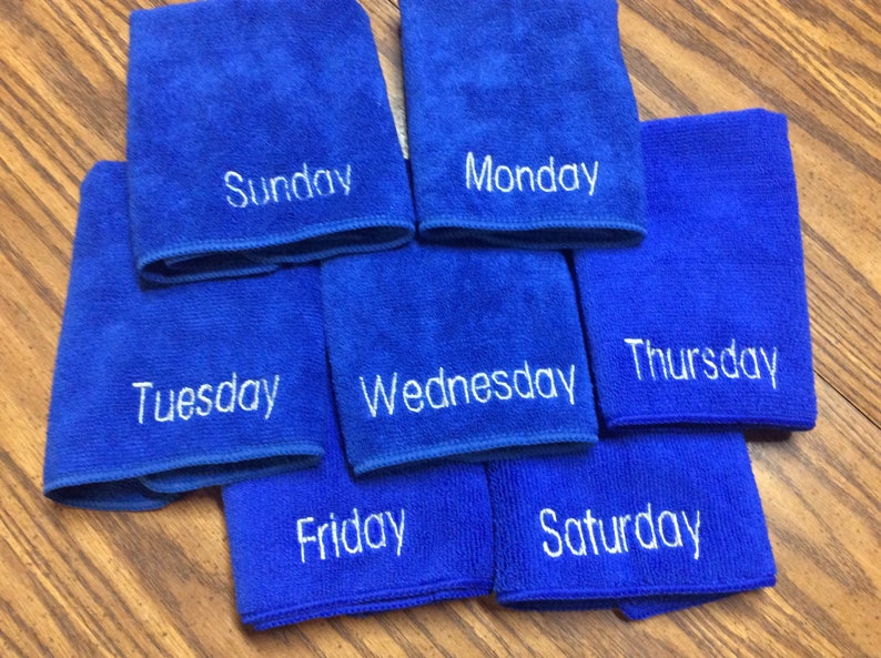 May include: Seven blue microfiber cloths, each embroidered with a day of the week in white. The cloths are arranged on a wooden surface. The days of the week are Sunday, Monday, Tuesday, Wednesday, Thursday, Friday, and Saturday.