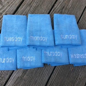May include: Eight light blue microfiber towels, each embroidered with a different day of the week in white. The towels are arranged on a weathered wooden surface.