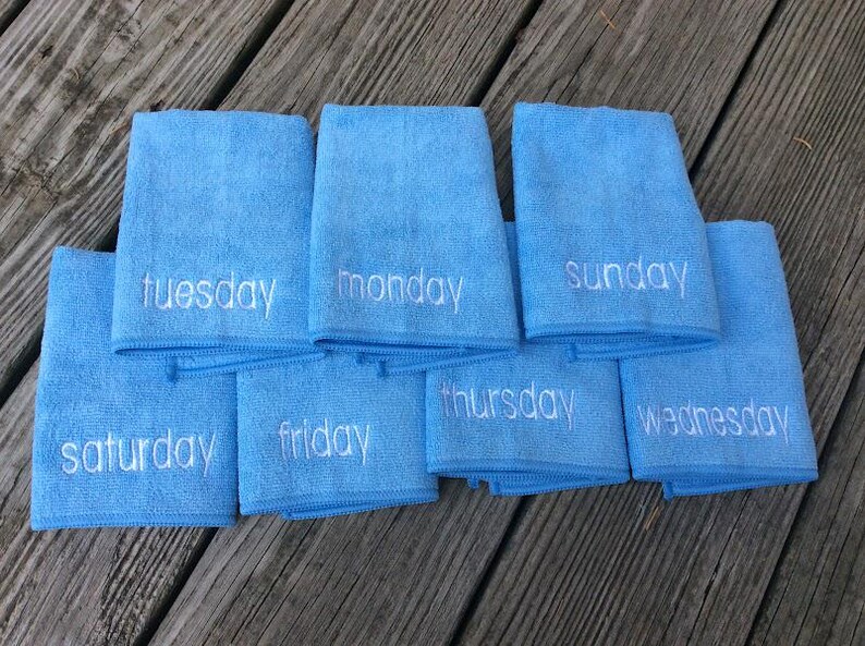 May include: Eight light blue microfiber towels, each embroidered with a different day of the week in white. The towels are arranged on a weathered wooden surface.