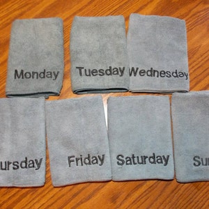 May include: Seven grey microfiber cloths, each embroidered with a day of the week in black: Monday, Tuesday, Wednesday, Thursday, Friday, Saturday, and Sunday. The cloths are arranged on a wooden surface.