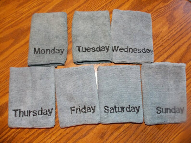 May include: Seven grey microfiber cloths, each embroidered with a day of the week in black: Monday, Tuesday, Wednesday, Thursday, Friday, Saturday, and Sunday. The cloths are arranged on a wooden surface.