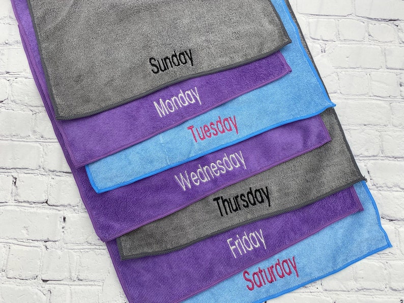 May include: A set of microfiber cleaning cloths in shades of grey, blue and purple, each embroidered with a day of the week: Sunday, Monday, Tuesday, Wednesday, Thursday, Friday, and Saturday. Ideal for cleaning.