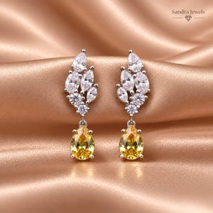 May include: A pair of silver-toned earrings featuring a cluster of clear, marquise and round-cut stones at the top, with a single yellow, pear-shaped drop stone at the bottom. The text Sandra Jewels is visible in the upper right corner.
