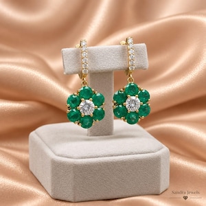 May include: A pair of gold drop earrings featuring a flower-shaped design. Each earring has six round green stones surrounding a central clear round stone. The gold hoop attachment is lined with small clear stones. Sandra Jewels text.