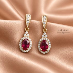 May include: A pair of gold drop earrings featuring oval-cut red rubies surrounded by halos of small white diamonds. The earrings have a triangular top section set with diamonds. The text Sandra Jewels is visible on the right side.