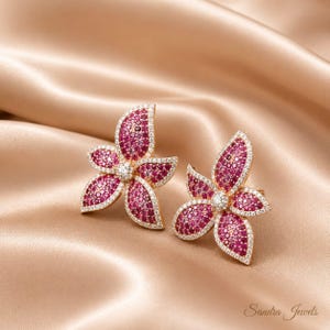 May include: A pair of flower-shaped stud earrings crafted in a gold-tone metal, featuring petals densely encrusted with small, vibrant pink stones and bordered by a delicate rim of tiny, clear white stones. The text Sandra Jewels is visible.