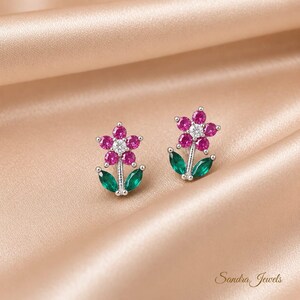 May include: A pair of silver stud earrings shaped like flowers, featuring five round pink stones surrounding a central clear stone, with two marquise-cut green stones forming the leaves at the base. The text Sandra Jewels is visible in the corner.