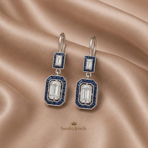 Puede incluir: Un par de pendientes colgantes con piedras transparentes de corte esmeralda rodeadas de piedras azul oscuro de corte baguette en un engaste plateado. Cada pendiente tiene una sección rectangular superior pequeña y una inferior grande.