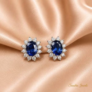 May include: A pair of stud earrings featuring a central oval-cut deep blue gemstone surrounded by a halo of small, clear, round-cut white stones set in a silver-toned metal. The text Sandra Jewels is visible in the bottom right corner.