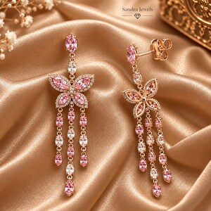 May include: A pair of rose gold-tone drop earrings featuring a butterfly motif adorned with marquise-cut pink and clear crystals. Each earring includes three dangling chains of alternating pink and clear stones. The text Sandra Jewels is visible.