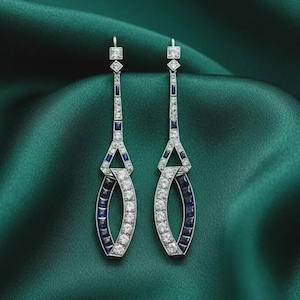 May include: A pair of long drop earrings featuring a geometric silver-toned metal frame set with clear white stones and deep blue rectangular stones. The design includes a vertical bar leading to an open oval shape with alternating stone accents.