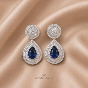 Op de afbeelding: Een paar zilverkleurige oorbellen met een cirkelvormig bovendeel en een peervormig onderste deel, bezet met heldere baguette- en ronde kristallen rondom een centrale diepblauwe peervormige steen. De tekst luidt SandraJewels.