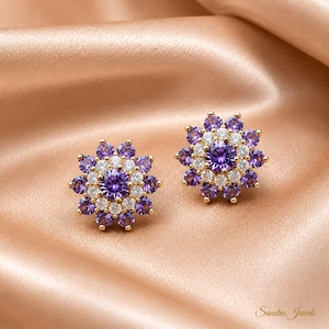 Könnte beinhalten: Ein Paar goldene Ohrstecker mit einem zentralen violetten Edelstein, umgeben von einem Kranz aus klaren weißen Steinen und einem äußeren Ring aus violetten Steinen in Blütenform. Der Text Sandra Jewels ist in der unteren Ecke zu sehen.