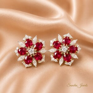 May include: A pair of floral-shaped earrings featuring oval-cut red rubies arranged in a petal pattern, accented with marquise and round-cut white diamonds set in gold. The text Sandra Jewels is visible in the bottom right corner.