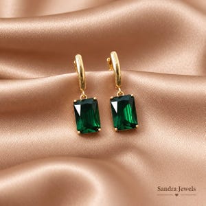 Op de afbeelding: Een paar goudkleurige oorbellen met rechthoekige, smaragdgroene gefacetteerde stenen die aan scharnierende ringen hangen. De sieraden worden getoond tegen een gladde, gedrapeerde stof. De tekst Sandra Jewels is zichtbaar in de rechterbenedenhoek.