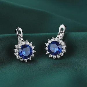 Può includere: Un paio di orecchini color argento con una grande pietra centrale rotonda blu intenso circondata da un alone di piccole pietre chiare e scintillanti. Ogni orecchino è dotato di una chiusura a leva incernierata e sicura.