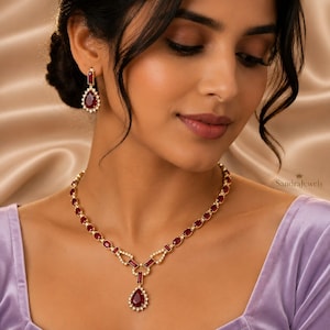 May include: A gold necklace and matching drop earrings set featuring deep red, pear-shaped and rectangular gemstones surrounded by small white stones. The necklace has a central pendant drop, and the earrings mirror the pear-shaped design.