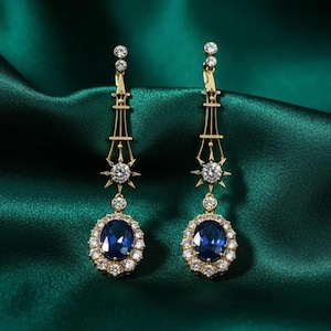 May include: A pair of gold drop earrings featuring an elongated, openwork architectural design. Each earring is accented with a small, round, clear faceted stone and terminates in a large, oval-cut deep blue stone surrounded by a halo of clear stones.