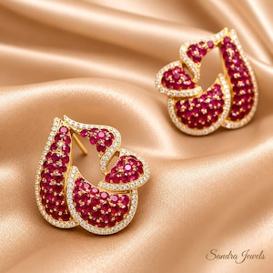 Könnte beinhalten: Ein Paar goldene Ohrringe mit tropfenförmigem Design, besetzt mit zahlreichen kleinen, leuchtend roten Steinen und umrandet von einem zarten Besatz aus winzigen, klaren weißen Steinen. Der Text Sandra Jewels ist unten rechts zu sehen.