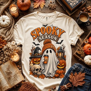 Może przedstawiać: Kremowy t-shirt z krótkim rękawem z nadrukiem graficznym i napisem "SPOOKY SEASON". Wzór przedstawia ducha w pomarańczowej czapce, trzymającego kubek kawy, w otoczeniu dyń, czaszki, latarni oraz nietoperzy.