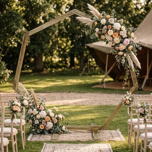 May include: A large wooden hexagonal frame structure decorated with floral arrangements featuring white, peach, and pink roses, greenery, and pampas grass. The frame is positioned on a patterned rug on the grass, with chairs placed on either side.