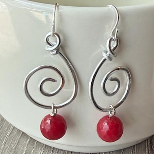 May include: A pair of silver-toned wire earrings featuring a spiral design with a single faceted red bead dangling from the bottom of each piece. The earrings are suspended from silver-toned ear wires and have a polished, reflective metallic finish.