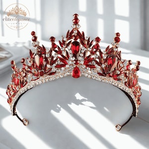 Puede incluir: Una tiara de oro rosa con cristales de color rojo intenso en forma de lágrima y talla marquesa dispuestos en un patrón puntiagudo y ornamentado. La base está bordeada por una hilera de pequeños diamantes de imitación blancos. Logo visible.