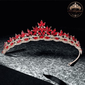 May include: A gold-toned tiara featuring a series of marquise-cut red crystals arranged in a floral pattern, accented with small clear rhinestones along the metal band. The piece has a curved, arched design with a central peak and side loops.