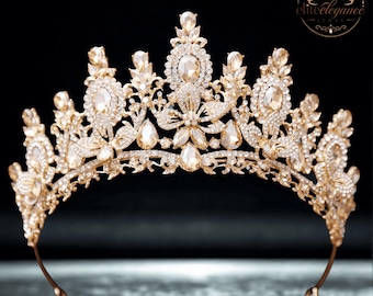 May include: A gold-toned tiara featuring an intricate metal filigree design adorned with numerous clear crystals and champagne-coloured teardrop-shaped stones. The crown displays a central floral motif and symmetrical patterns throughout its structure.