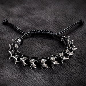 May include: A black cord bracelet featuring a series of metallic, silver-toned vertebrae-shaped beads. The bracelet includes an adjustable sliding knot closure with two thin black cords extending from the centre, creating a secure fit.