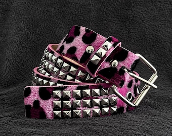 May include: A belt featuring a pink and black leopard print pattern on a textured surface. The belt is adorned with three horizontal rows of silver-colored metal pyramid studs and includes a silver-colored metal buckle and matching hardware.