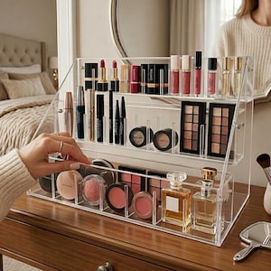 May include: A clear acrylic tiered makeup organizer holding various cosmetics including lipsticks, eyeshadow palettes, blush compacts, and perfume bottles. The organizer features multiple shelves and dividers to keep beauty products upright and visible.
