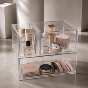 May include: A clear acrylic makeup organizer with two top compartments and one wide bottom drawer. The organizer holds various skincare and cosmetic products, including bottles labeled SK-II, The Ordinary, Chanel, and a palette labeled NUDE.