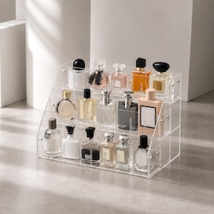 Peut inclure: Un présentoir à étages en acrylique transparent contenant quatorze flacons de parfum en verre de formes et de tailles variées. L'organisateur comporte trois niveaux étagés, permettant une disposition ordonnée des contenants de parfum.