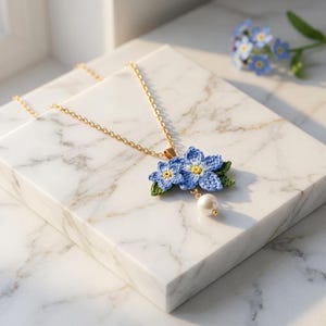 May include: A gold-tone chain necklace featuring a pendant of two crocheted light blue forget-me-not flowers with yellow centers and green leaves. A single white pearl bead hangs from the base of the floral pendant, adding a delicate accent.