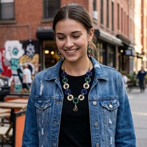 May include: A necklace featuring alternating circular links in shades of blue, green, and cream, finished with a single teardrop-shaped pendant. The piece is worn over a black top and a distressed blue denim jacket with silver-toned metal buttons.