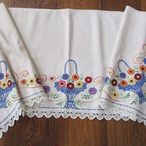 May include: Cream-colored embroidered table runner with a floral basket design. The baskets are blue, filled with colorful flowers. The edges are trimmed with white lace and blue stitching.