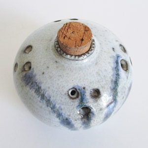 May include: A handmade ceramic bottle with a cork stopper. The bottle is round with a speckled white glaze and blue streaks. It has several small holes around the sides and top. The cork is a light brown color.