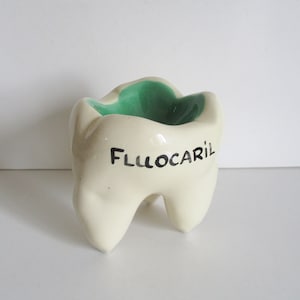 May include: A ceramic tooth-shaped container with a cream-colored exterior and a green interior. The word "FLUOCARIL" is written in black on the front of the tooth. The container has three root-like structures at the base.